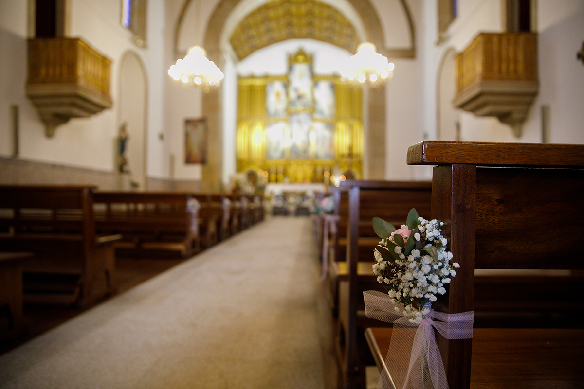 Um plano detalhado revela um ramo de flores brancas preso com uma fita cor-de-rosa a um banco da igreja, um gesto delicado e simbólico que anuncia a beleza do momento prestes a acontecer.