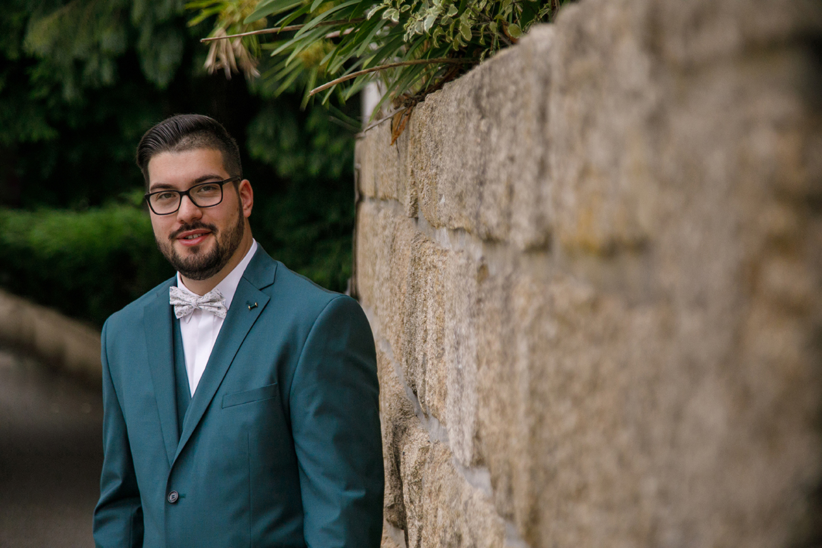 Quase encostado a um muro de pedra, Tiago sorri para a câmara com uma mistura de seriedade e doçura, enquanto o fundo desfocado destaca a sua presença calma e confiante, como se guardasse um segredo de amor.