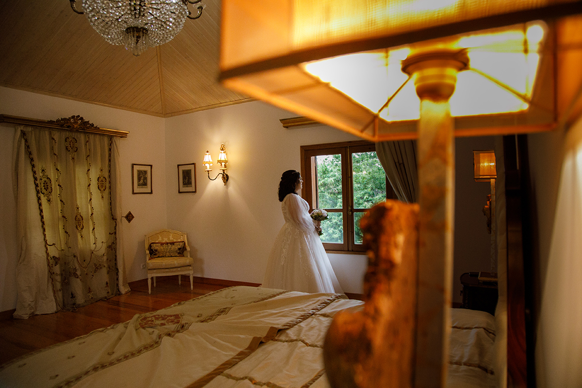 De costas para a câmara, Carolina contempla a luz que invade o quarto pela janela, o bouquet firme na mão, num momento de silêncio onde as emoções se entrelaçam com a calma, o candeeiro ao lado acende um brilho cálido que parece abençoar a noiva.