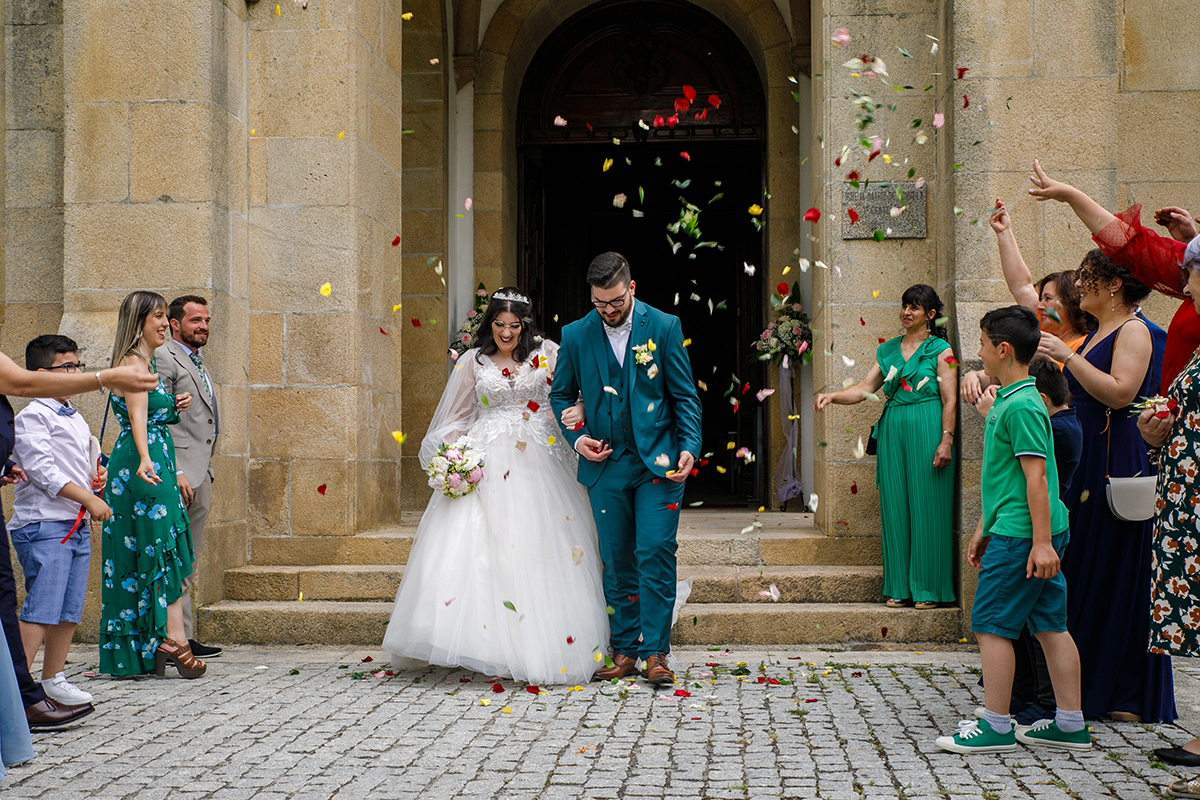Carolina e Tiago saem da igreja rodeados pelos convidados que atiram pétalas de flores, eles abaixam as cabeças num gesto terno, os rostos iluminados por sorrisos felizes e olhos brilhantes de emoção.