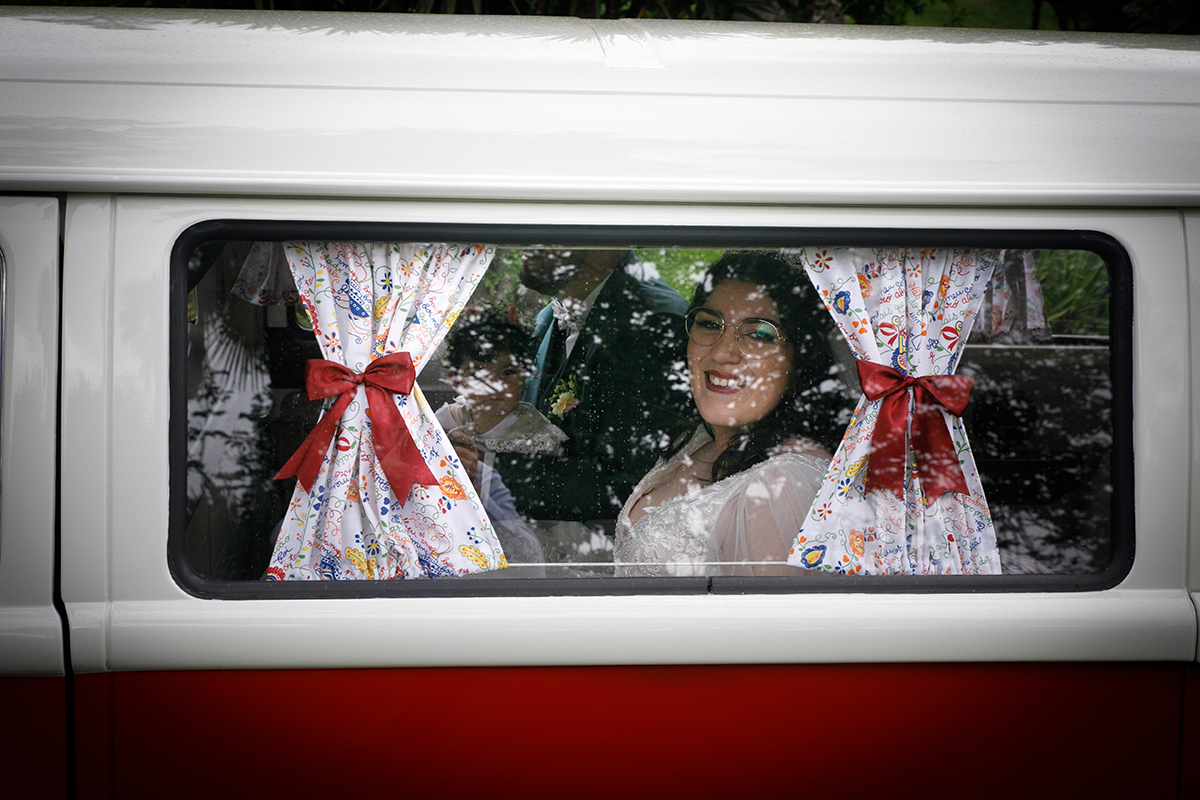 O vidro da VW Pão de Forma revela a noiva ao pé da janela a sorrir para o exterior, enquanto o noivo do outro lado da carrinha, envoltos pelas cortinas decoradas com lacinhos vermelhos que lhes conferem um charme vintage e íntimo.