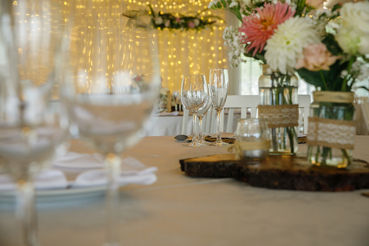 A mesa do restaurante espera pelos convidados com uma decoração perfeita de flores brancas e rosas, a cortina branca com luzes amarelas ao fundo cria uma atmosfera suave e acolhedora, pronta para celebrar o amor.