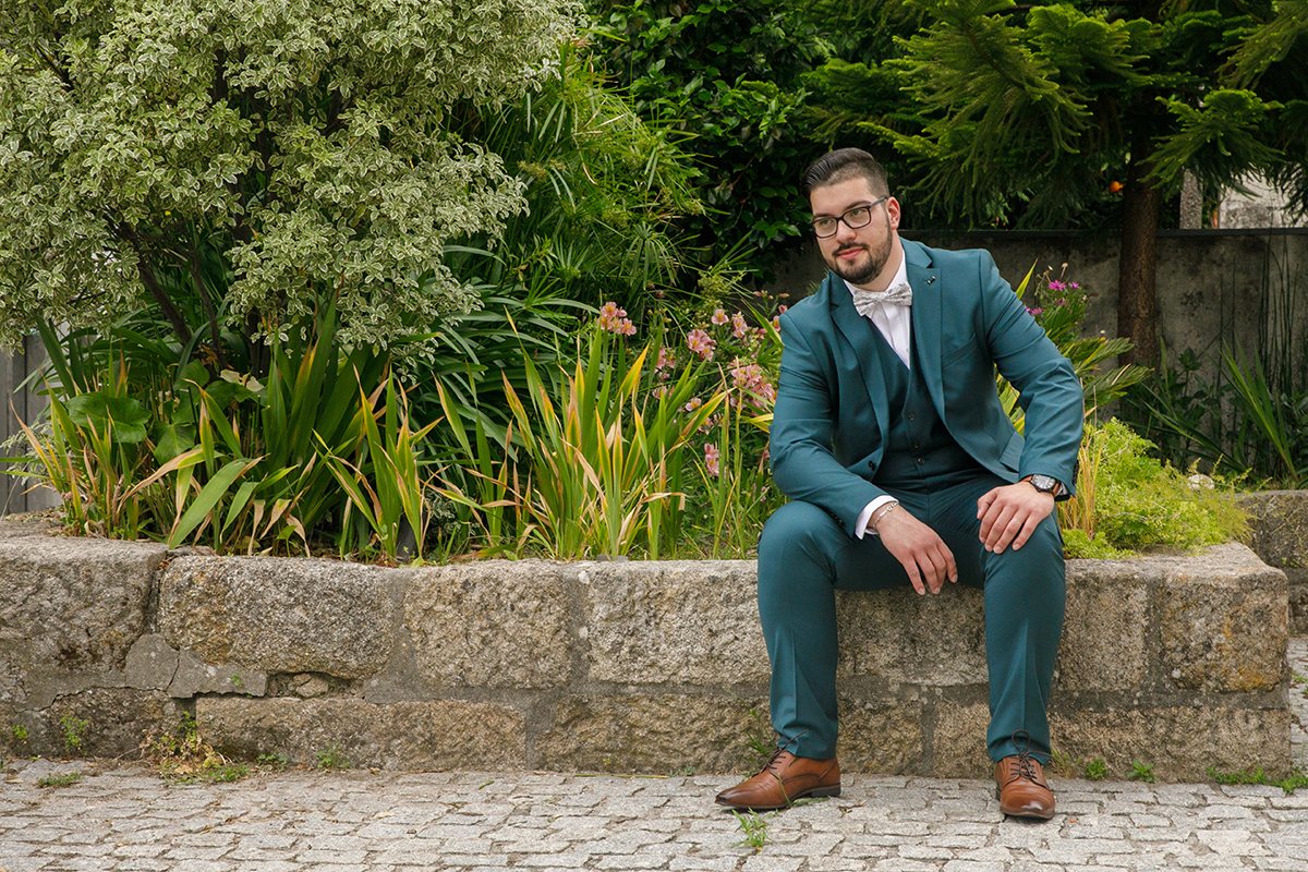 Sentado num pequeno muro de pedra, Tiago repousa os braços sobre as pernas e dirige um olhar feliz para o lado, envolto pelo verde das plantas, flores e árvores que pintam a cena com a promessa de um novo começo.