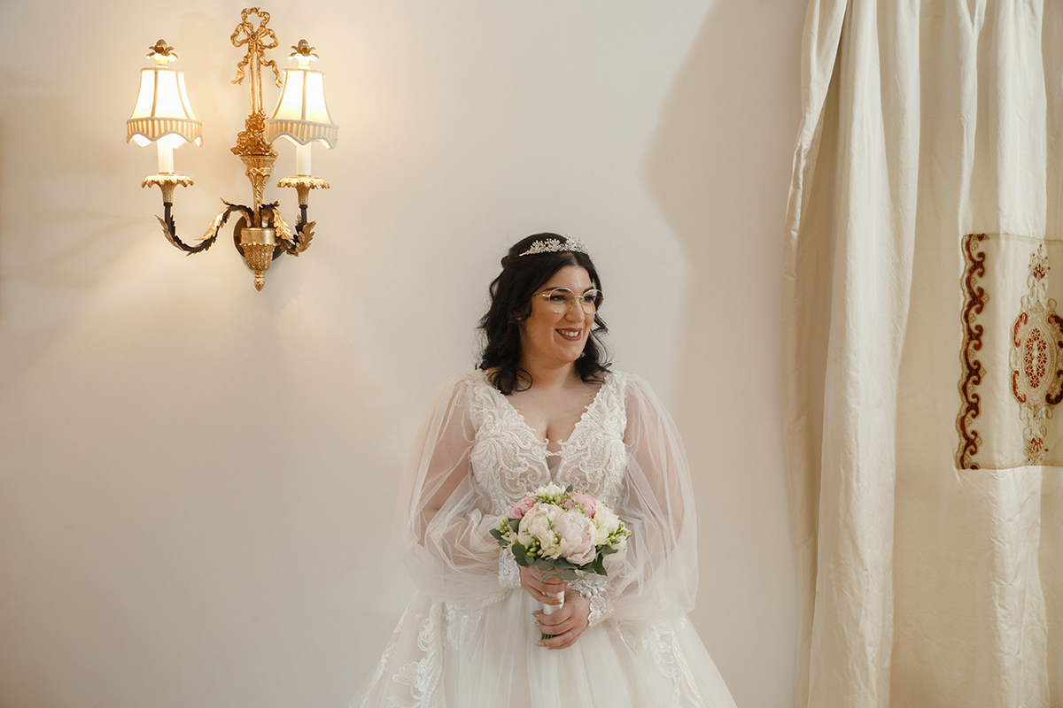 Carolina ocupa o centro da imagem, segurando o seu bouquet com ternura, o vestido de encantar espalha-se em delicados detalhes e a cortina bege ao lado molda suavemente a cena, como se cada elemento sussurrasse um conto de amor.