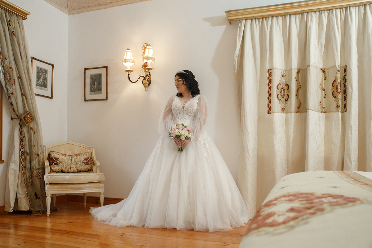Frente a uma parede branca, Carolina dirige o olhar pensativo para a janela, o sorriso leve e o bouquet em mãos fazem sobressair a delicadeza do vestido, que acompanha a silhueta esguia da noiva com elegância.