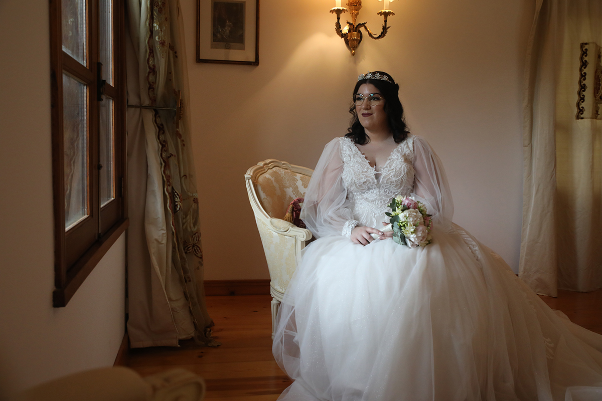 Sentada num cadeirão bege, Carolina contempla a janela com uma expressão serena e feliz, o bouquet repousa delicadamente nas pernas, num retrato de paz e beleza genuína.