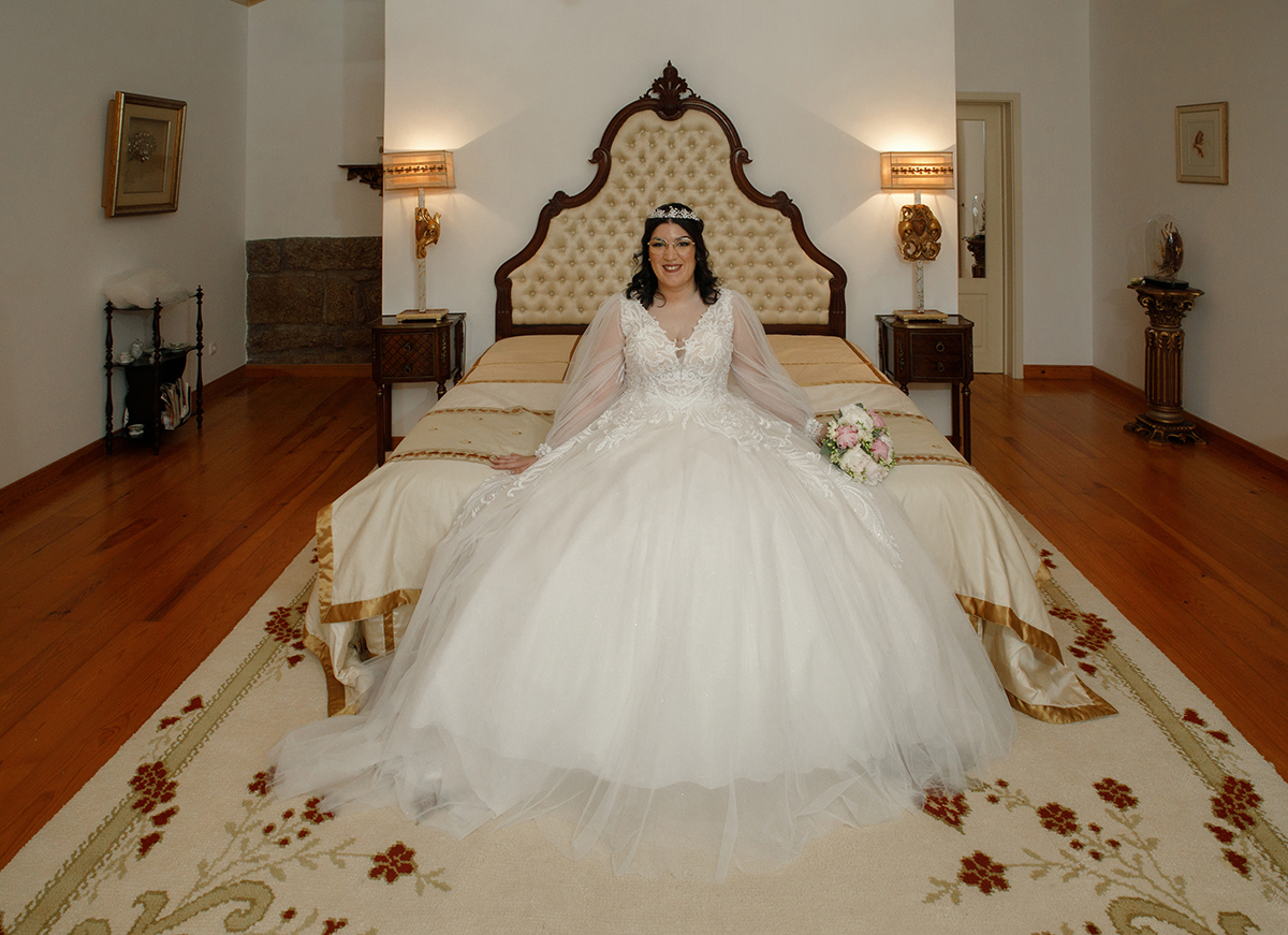 Sentada ao fundo da cama, Carolina olha para a câmara com um sorriso de felicidade genuína, as mãos repousadas suavemente sobre a cama junto ao bouquet, numa imagem que transpira aconchego e sonho.