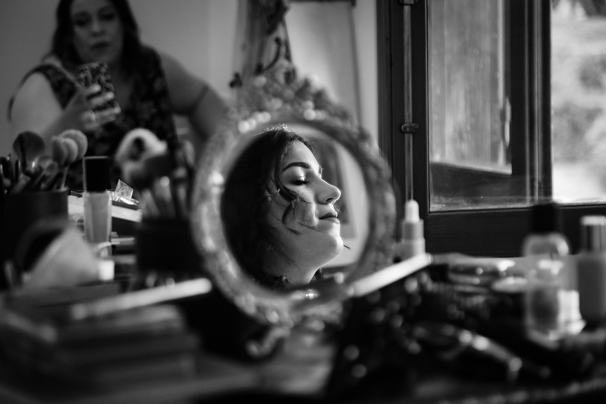 Em preto e branco, a penteadeira antiga destaca um espelho redondo onde o reflexo de Carolina surge sereno, os olhos fechados num sorriso leve, enquanto uma amiga capturava este momento intemporal ao fundo, envolvido em sombras e luz.