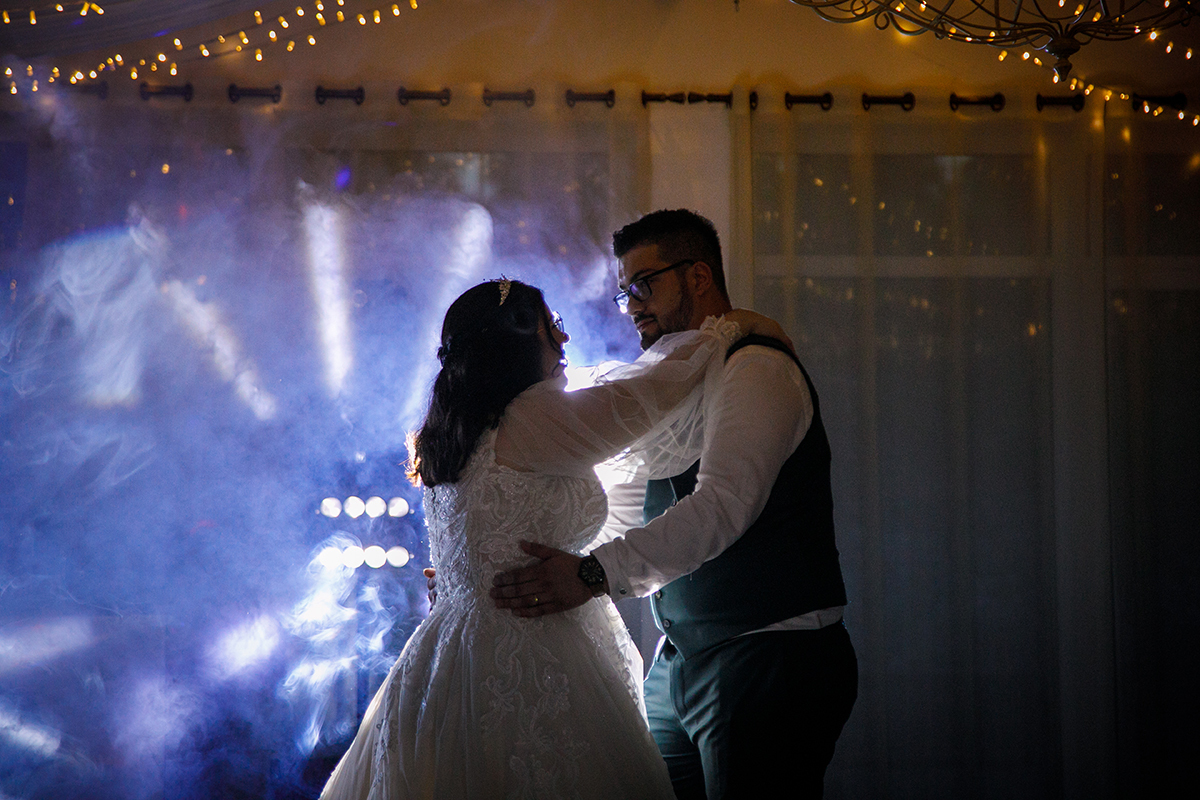 Envoltos numa atmosfera de luz branca e amarelas cintilantes na parede, Carolina e Tiago olham-se nos olhos, abraçados e felizes, Tiago segura a cintura dela enquanto ela repousa as mãos nos ombros dele, um retrato de cumplicidade profunda.