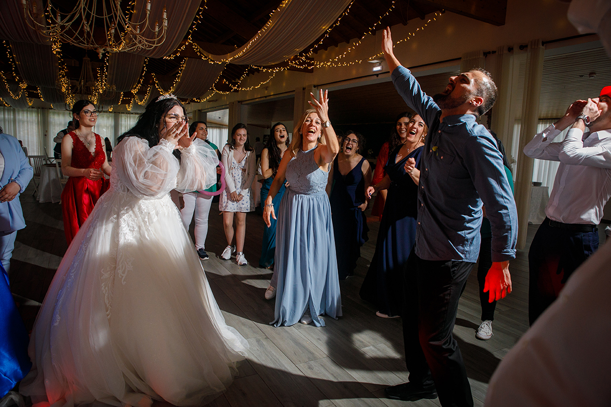 A noiva, de mãos junto à boca, expressa uma alegria contagiante enquanto um convidado à frente grita em êxtase com o braço erguido, ao redor a multidão ri, canta e celebra, enchendo o ambiente de energia e felicidade.