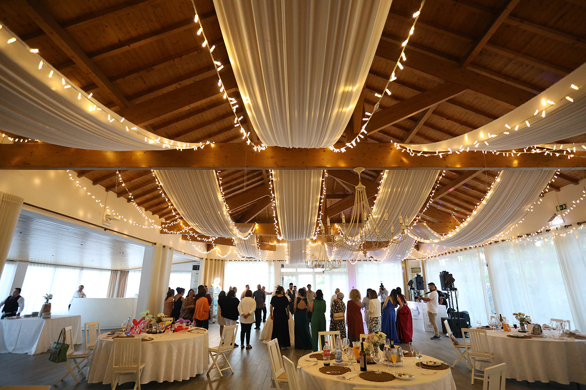 A sala de receção revela um espaço encantador com tecidos brancos pendendo do teto e luzes delicadas que criam uma atmosfera mágica, as mesas organizadas aguardam os convidados para celebrarem em conjunto este dia tão especial.