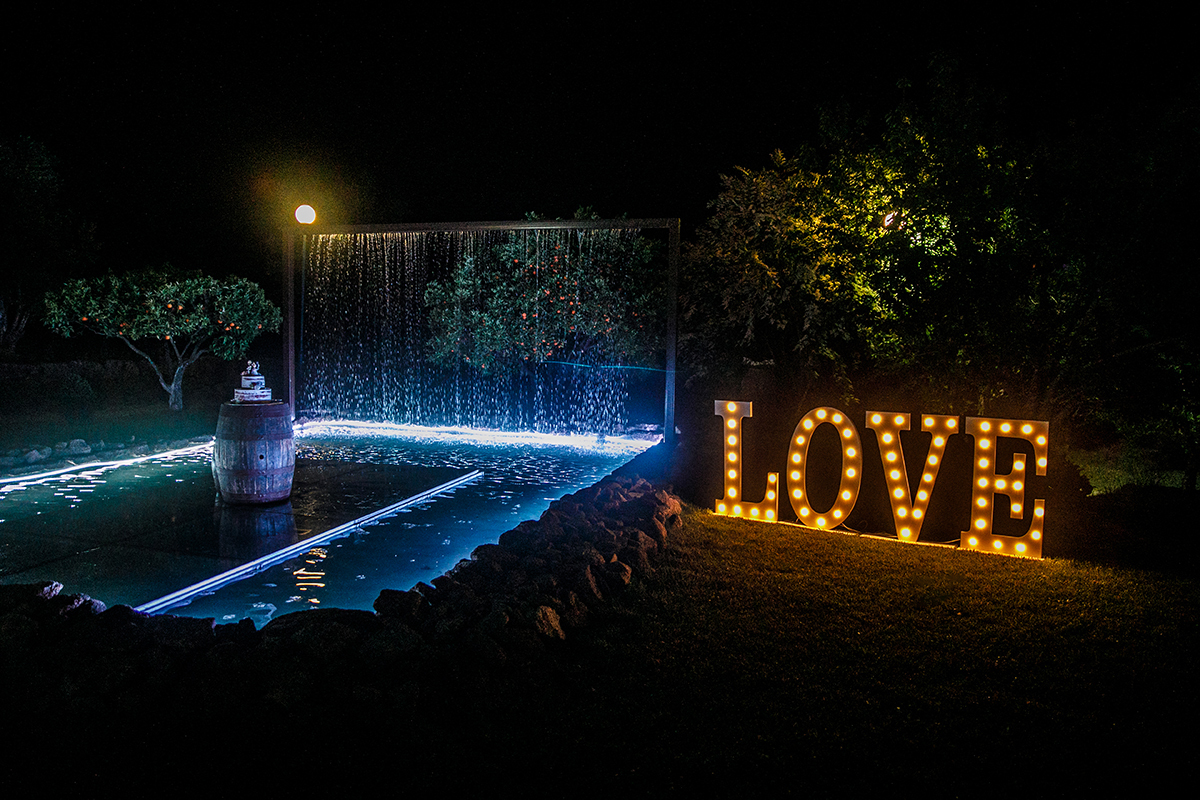 Num cenário de sonho, um lago ou piscina com bordas iluminadas acolhe uma plataforma onde repousa o bolo segurado por uma pipa de vinho, enquanto fios de água finos caem suavemente de uma estrutura de madeira e as letras 