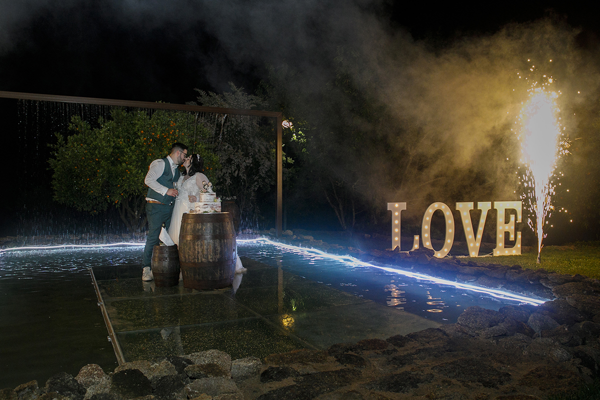 Os pombinhos partilham um beijo intenso sobre a plataforma ao lado do bolo e copos de champanhe, enquanto uma garrafa lança um fogo de artifício, celebrando o amor com brilho e emoção numa imagem que respira felicidade.