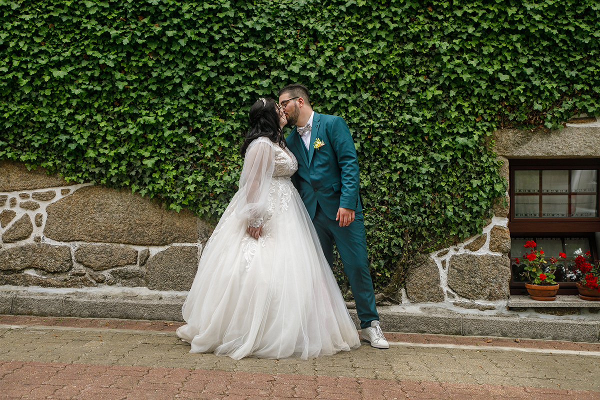 Ao pé da parede de pedra coberta por trepadeiras, eles trocam um beijo apaixonado que parece fundir dois corações num só, enquanto a textura da pedra acrescenta força e naturalidade a este instante de ternura.