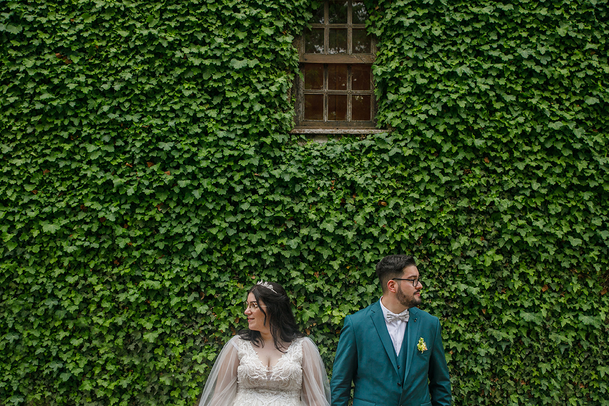 Ambos no centro da foto, lado a lado, virados para direções opostas mas com a felicidade estampada no rosto, contra uma parede revestida por trepadeiras e uma janela de madeira que acrescenta textura e charme à composição.