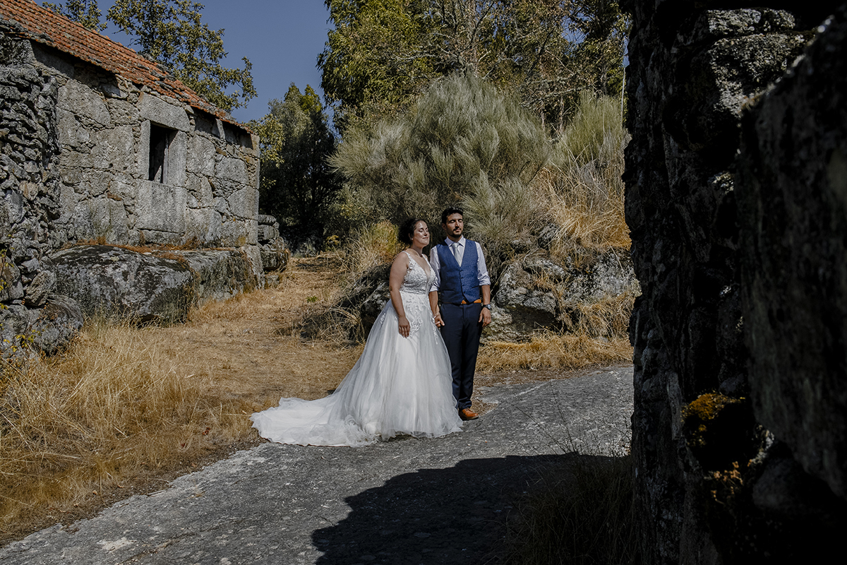 À direita da foto, uma parede de pedra antiga contrasta com o casal no centro, lado a lado, olhos fechados, banhados pela luz do sol, transmitindo paz e introspecção.