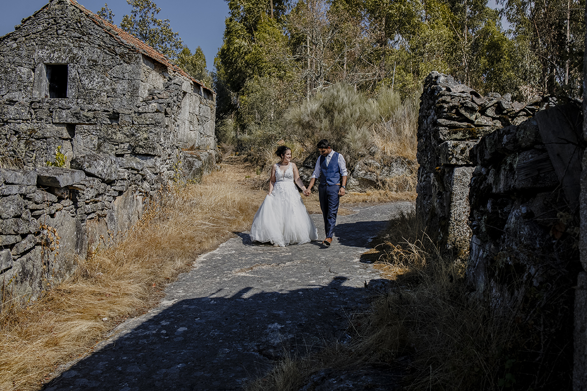 De corpo inteiro, o casal caminha de mãos dadas em direção à câmara, Miguel olhando para o chão e Juliana sorrindo para ele, com casas antigas compondo o cenário.