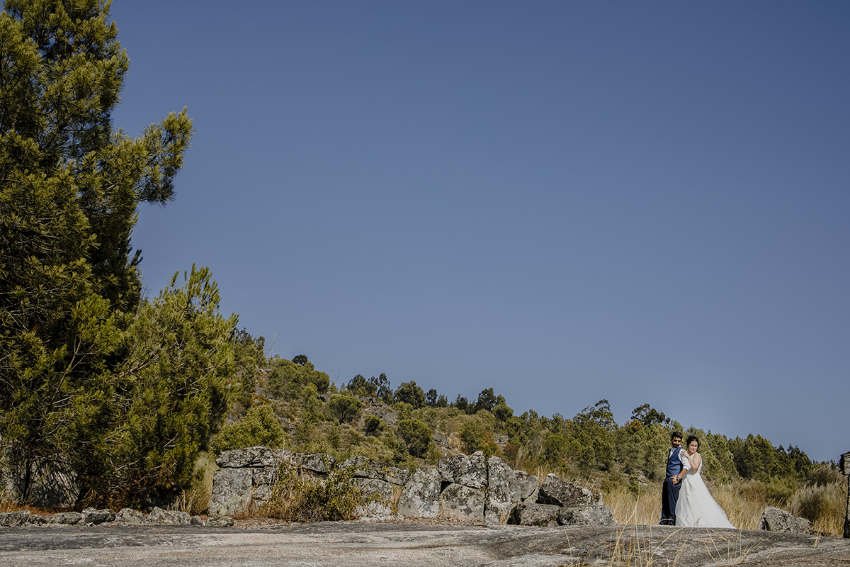 À direita da imagem, Juliana está atrás de Miguel, mãos nos seus ombros, ambos olhando para o chão, envoltos por um cenário de rochas e vegetação, simbolizando apoio e introspecção.