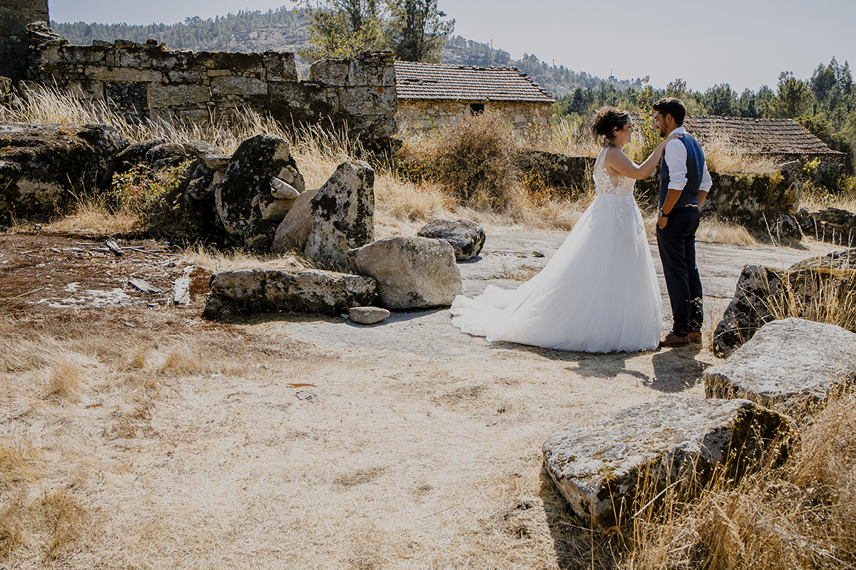 Entre pequenas rochas, Juliana e Miguel estão de frente um para o outro, ela com a mão no ombro dele, ambos sorrindo, capturando a alegria do momento.