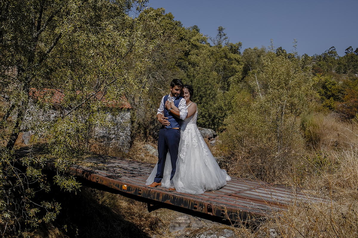 Ainda na ponte, de corpo inteiro, Juliana está atrás de Miguel, mãos no seu peito e barriga, enquanto ele segura as mãos dela, num gesto de união e carinho.