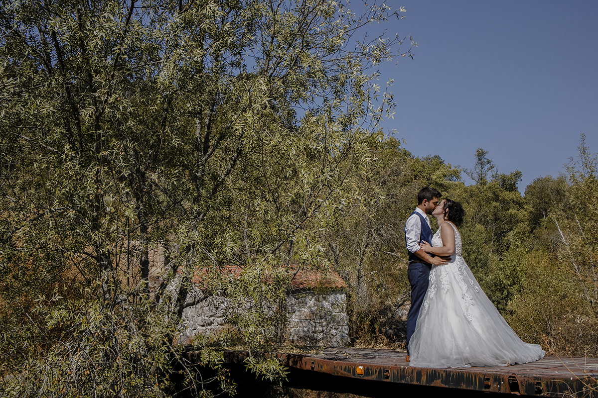 À direita da imagem, o casal se abraça pela cintura, frente a frente, com uma árvore verde à esquerda e uma construção antiga visível entre os ramos ao fundo.