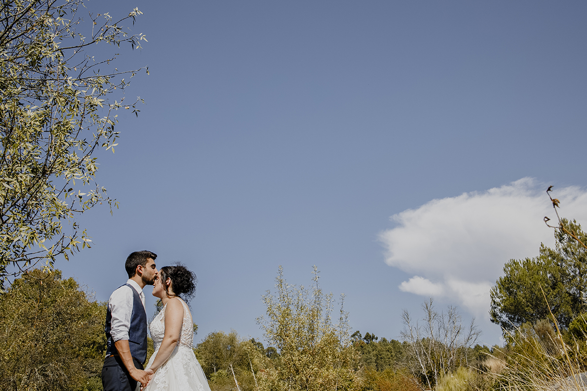 Da cintura para cima, à esquerda da foto, Miguel beija a testa de Juliana enquanto dão as mãos, com uma paisagem verde e o céu ao fundo, simbolizando ternura e proteção.