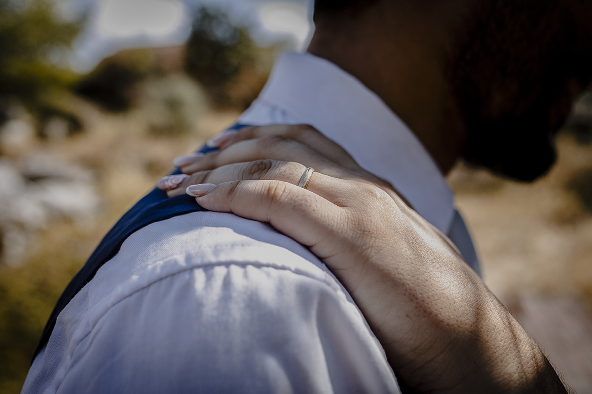Um close-up do ombro de Miguel até seus lábios, destacando a mão de Juliana sobre seu ombro, com a aliança em evidência, simbolizando o compromisso entre eles.
