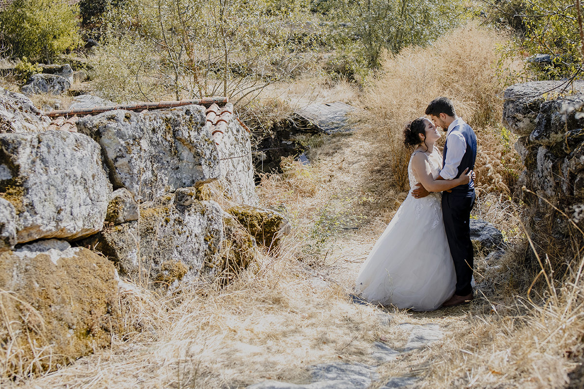Entre muros de pedra antigos, à direita da foto, o casal está de perfil, frente a frente, abraçados pela cintura, encostando os narizes com olhos fechados, em serenidade.