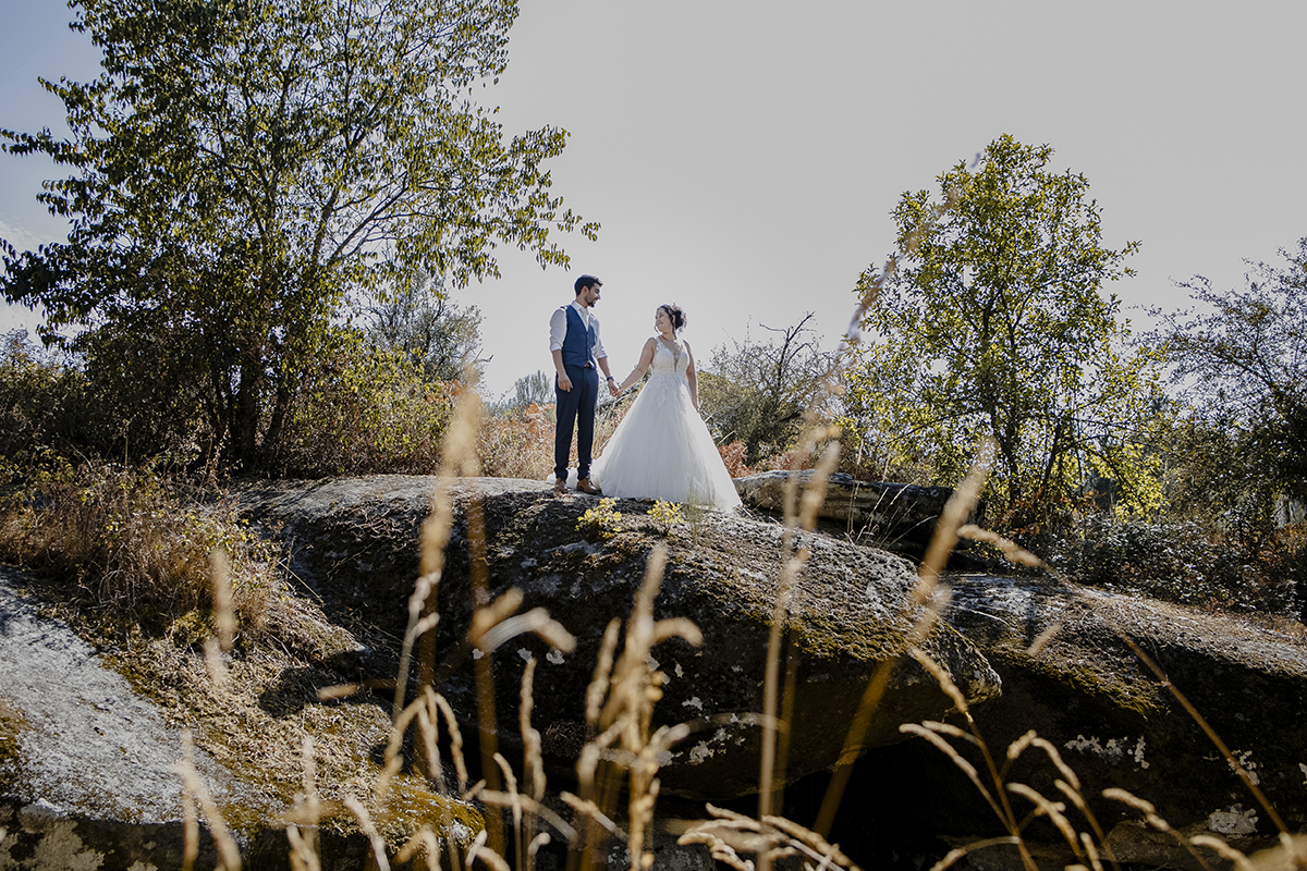Num plano mais afastado, de corpo inteiro, o casal está lado a lado de mãos dadas sobre uma rocha, olhando um para o outro com sorrisos, com plantas castanhas em primeiro plano.