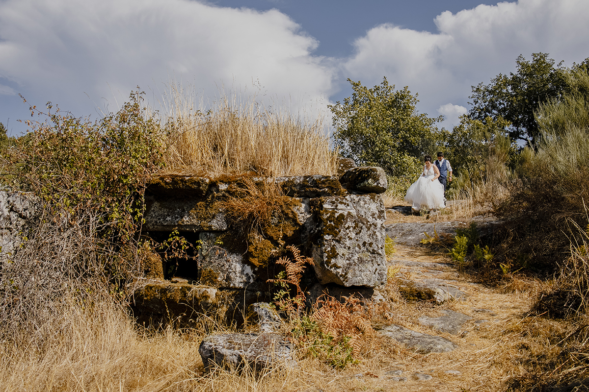 Ao ar livre, descem por um caminho, Juliana à frente e Miguel logo atrás, ambos olhando para o chão felizes, com um muro antigo à esquerda da foto.