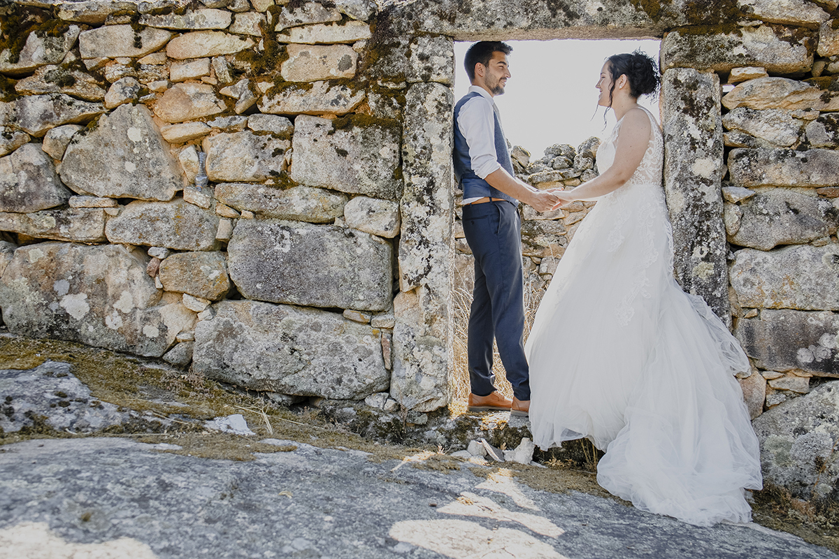 Diante de uma parede de uma casa antiga com um buraco em forma de porta, o casal está lá dentro, encostados lado a lado, frente a frente, de mãos dadas, encarando-se felizes.