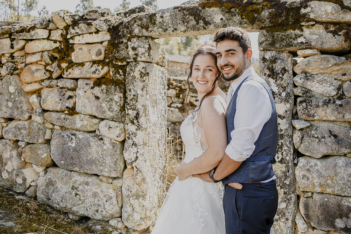 À frente da mesma porta, de lado para a câmara, Miguel abraça Juliana pela cintura enquanto ela coloca as mãos sobre a barriga, ambos olhando para a câmara com sorrisos.
