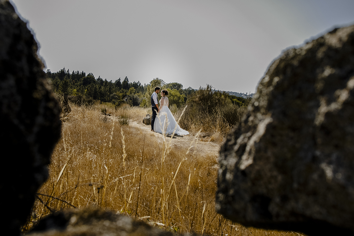 Ao fundo, frente a frente, de mãos dadas, encarando-se serenamente, vistos através de um buraco entre pedras, com pedras ao redor da lente da câmara.