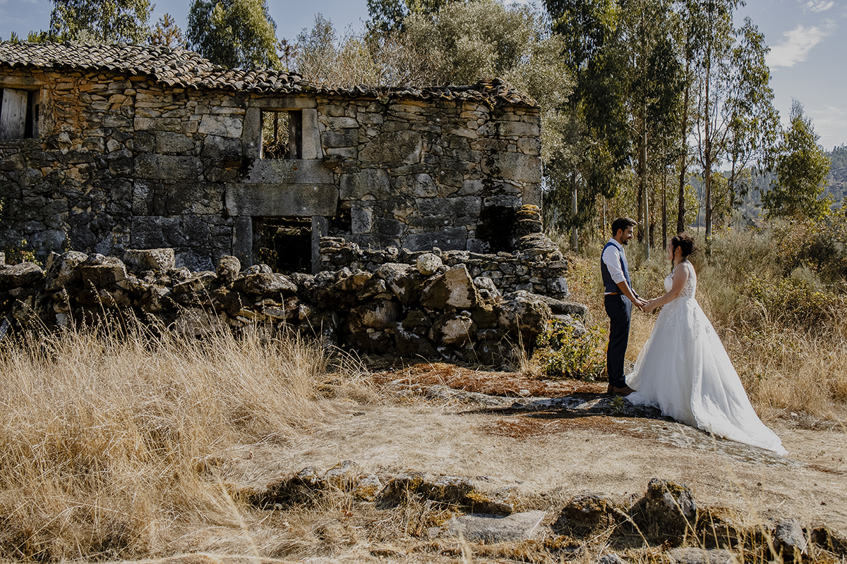 Diante de uma casa e muro antigos, de corpo inteiro, à direita da foto, frente a frente, de mãos dadas, encarando-se e sorrindo, simbolizando o amor duradouro.