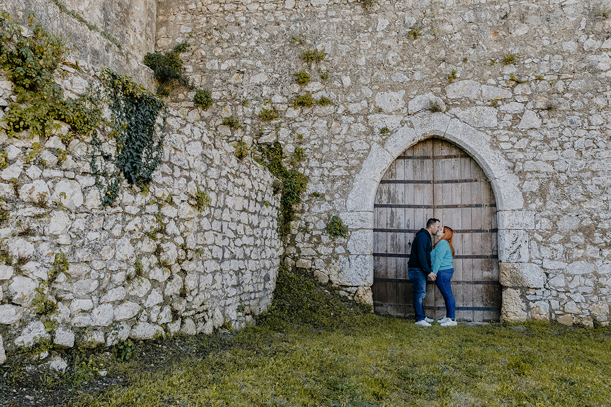 Ao fundo do portão do castelo, envoltos pelo tempo e pela arquitetura, os dois beijam-se de mãos dadas. As trepadeiras acompanham o momento como flores que desabrocham para o amor.