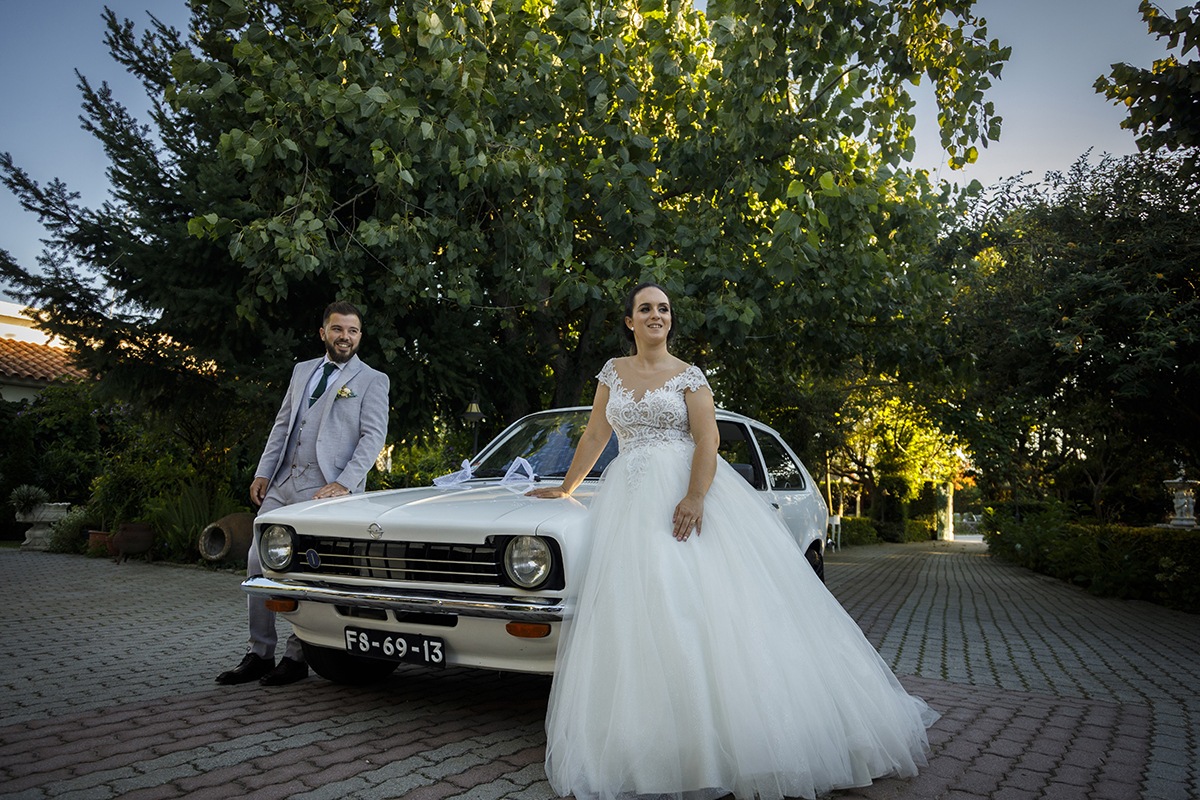 Descrição: Aqui com um Opel antigo no centro da foto vemos os dois cada um de cada lado do carro os dois estão virados para a frente para a câmara e estão de corpo inteiro e felizes a sorrirem ligeiramente para o lado.
