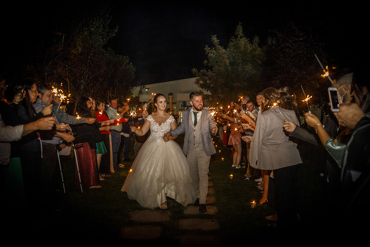 Descrição: Aqui já é noite e os dois caminham por um corredor de convidados com palitos de fogo eles caminham de mãos dadas felizes a olharem para os convidados.