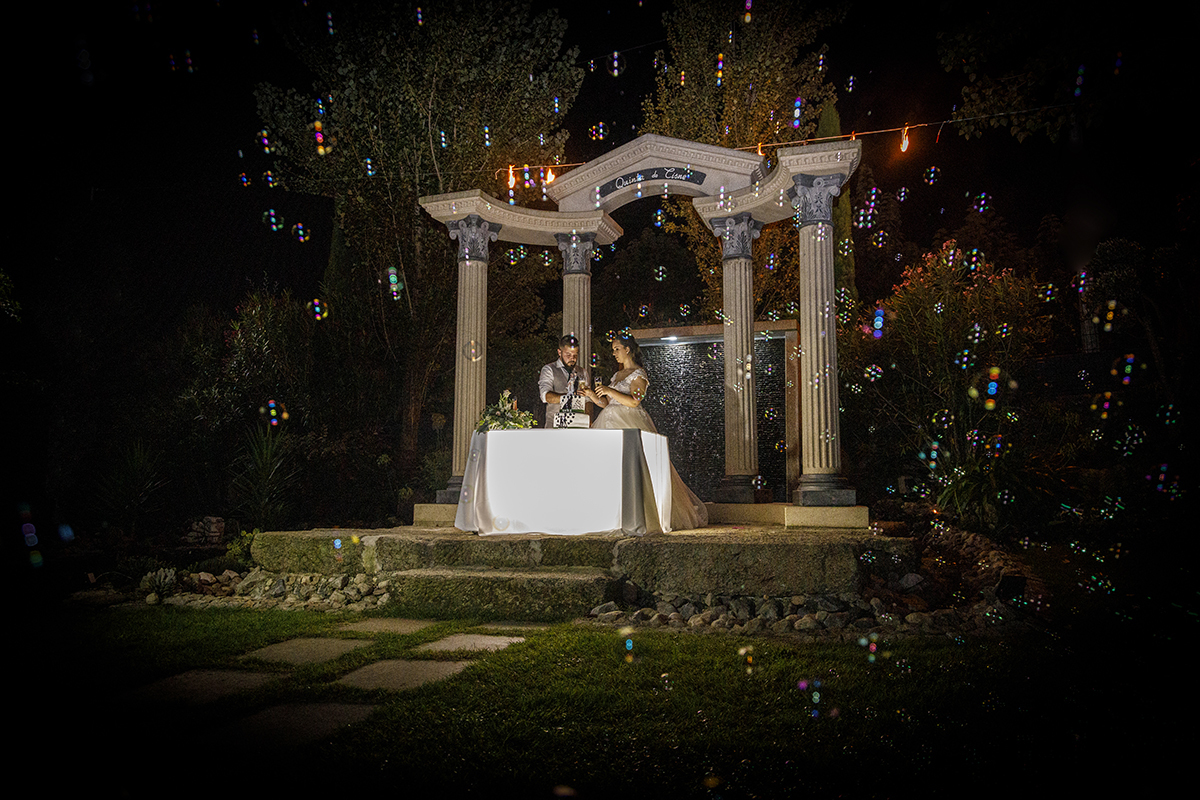 Descrição: Aqui com bolas de sabão pelo ar vemos os dois num cenário rústico com pilares e á frente uma mesa que tem o seu bolo e onde eles estão com os seus copos de champanhe na mão a brindarem.