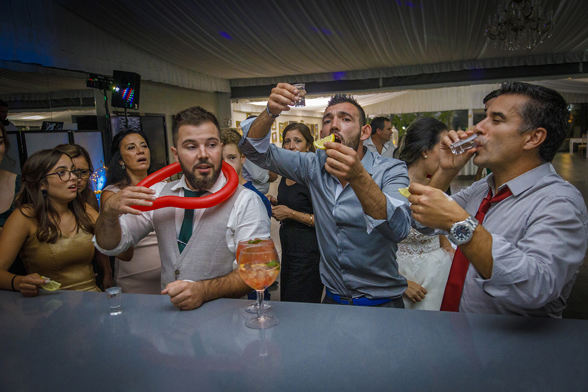 Descrição: Aqui vemos em um clima de festa o noivo com um balão vermelho enrolado ao pescoço e ao seu lado estão os convidados a tomarem shots.