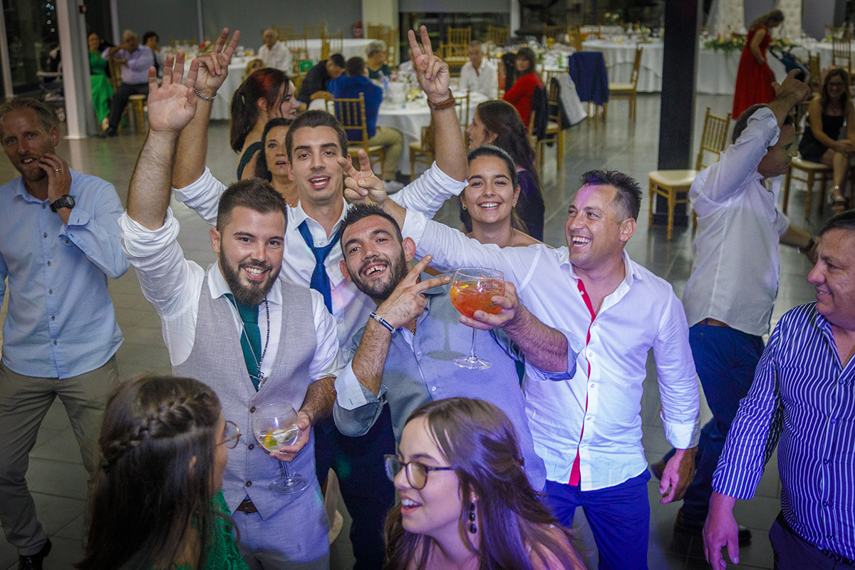 Descrição: Aqui temos uma foto com vinheta e vemos o noivo e os convidados com as mãos no ar copos na outra mão e sorrisos na cara.