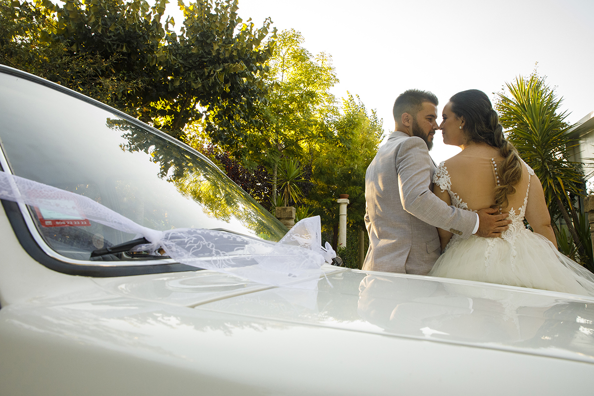 Descrição: Aqui vemos os dois encostados á parte lateral dianteira do Opel antigo e os dois estão lado a lado de costas e ele coloca a sua mão nas costas dela e os dois viram a cara um para o outro e encostam os seus narizes.