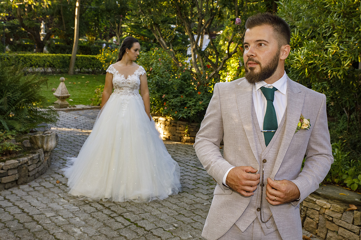 Descrição: Aqui sobre um caminho de paralelos vemos a Marta atrás de corpo inteiro de frente mas virada para o lado para baixo e depois á frente temos Ricardo da cintura para cima e ele olha ligeiramente para o lado para cima sério e com as mãos no casaco