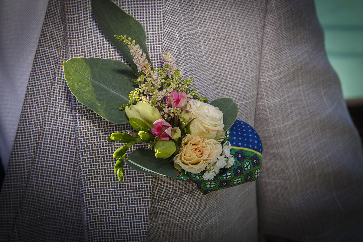 Aqui o olhar mergulha num plano de pormenor com vinheta onde o bolso do casaco exibe com elegância um arranjo delicado e um lenço colorido que contrasta com o cinzento subtil do tecido riscado, num detalhe de charme absoluto.