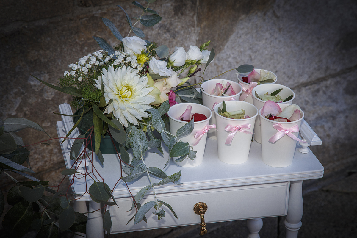 Descrição: Aqui vemos em uma foto com vinheta uma mesinha com um arranjo de flores e com uns copinhos com um laço cor de rosa e algo por dentro que não consigo identificar o que é 