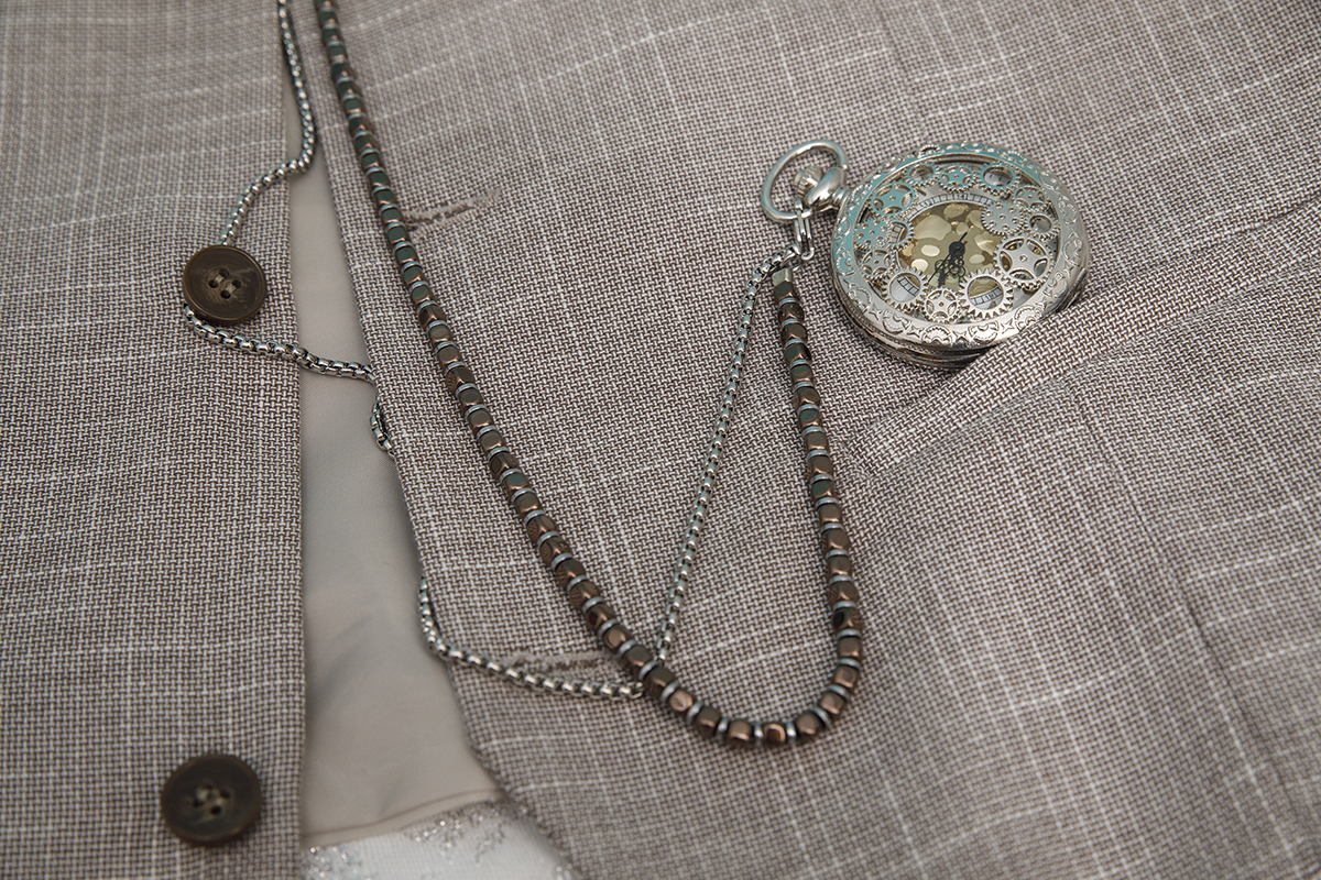 Os olhos descansam agora num colete estendido onde os botões castanhos e um antigo relógio de bolso nos contam histórias de herança e de estilo, num plano de detalhe cheio de personalidade.