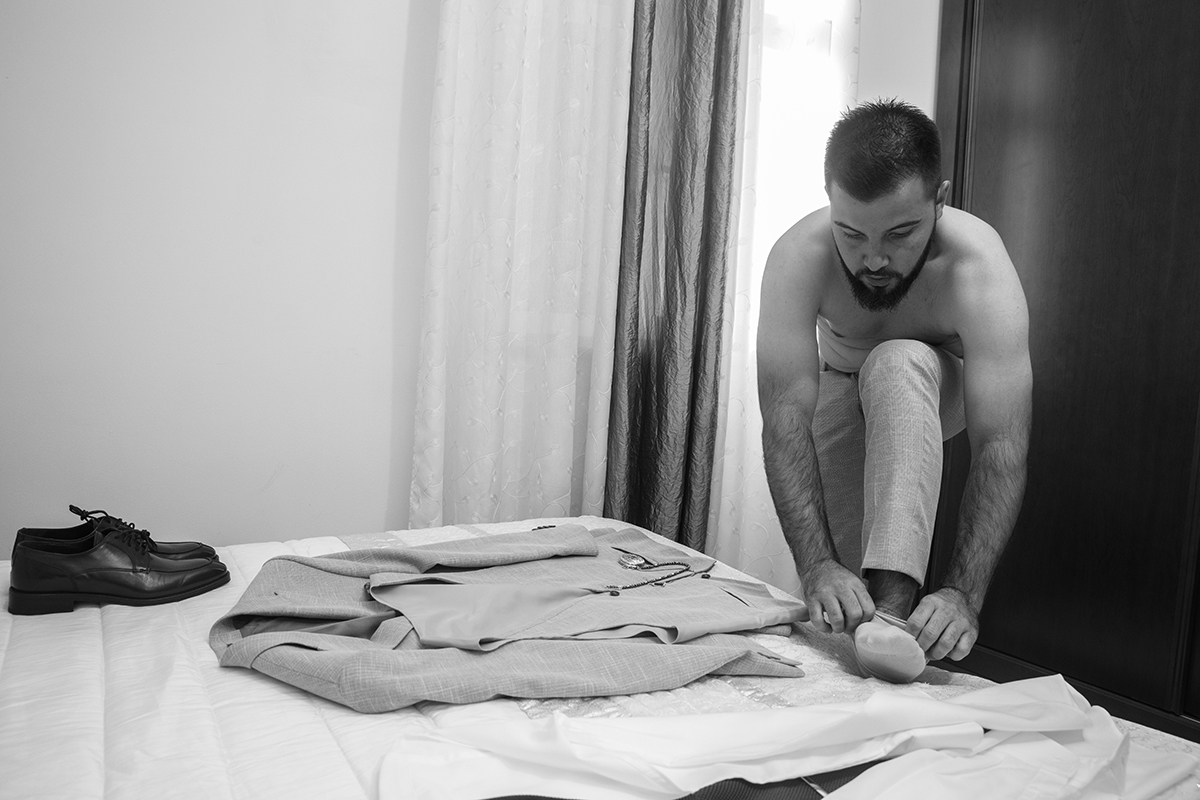 Ainda imerso na poesia do preto e branco, o fato repousa sobre a cama como quem aguarda o seu momento enquanto Ricardo, com uma perna apoiada, inclina-se sereno para calçar as meias, num gesto cheio de quietude e intenção.