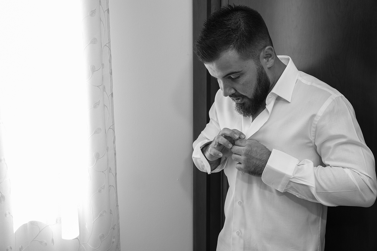 Num plano mais próximo, da cintura para cima, vemos Ricardo junto à janela, a luz suave a contornar-lhe o rosto enquanto ele aperta os botões da camisa com calma, num gesto atento que parece suspenso no tempo.