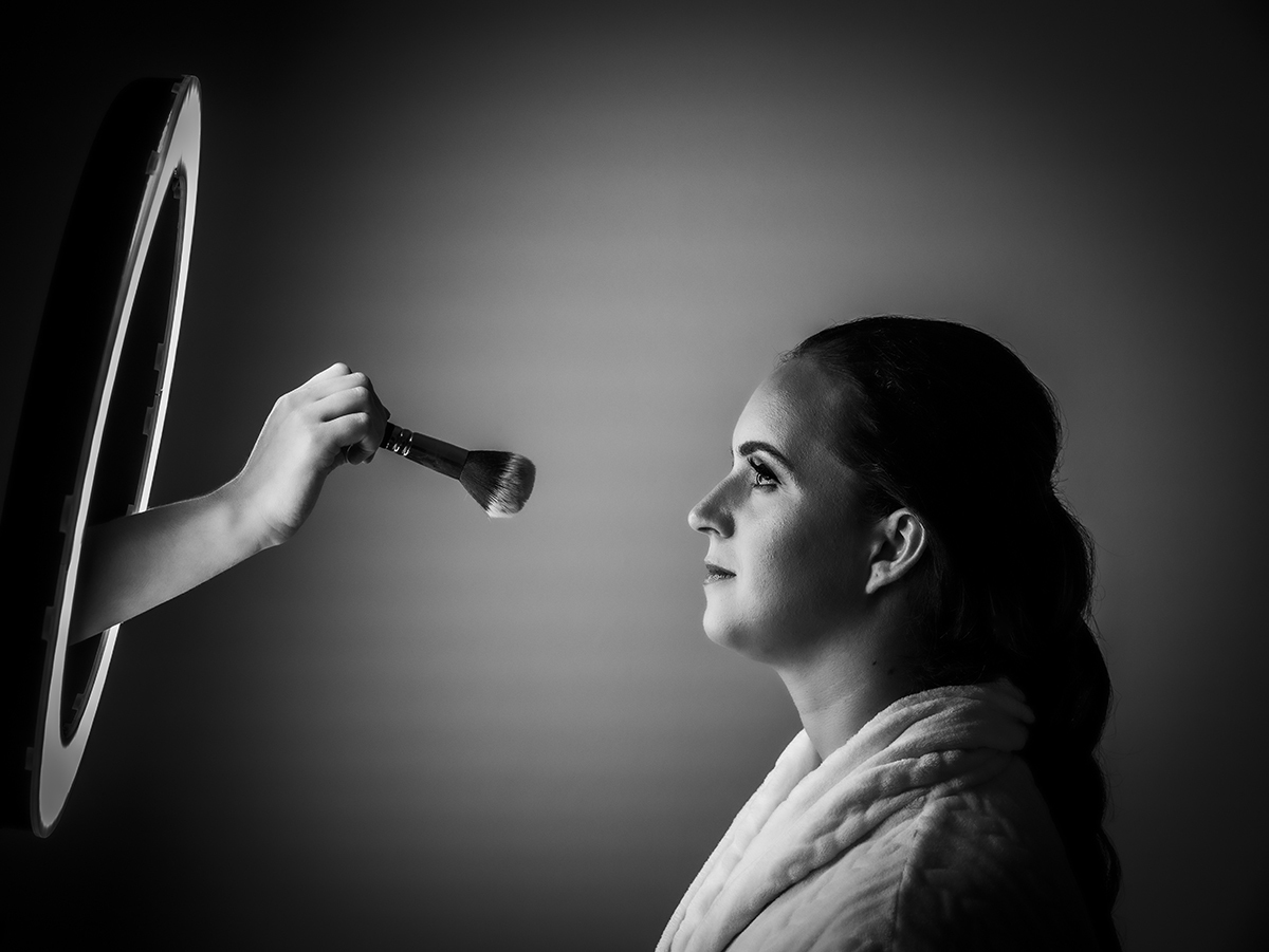 Num jogo de luz e composição, a mão da maquilhadora surge de dentro do ring light com um pincel suspenso diante de Marta que, vista de perfil, olha para ele séria e feliz, como se o momento a esculpisse.