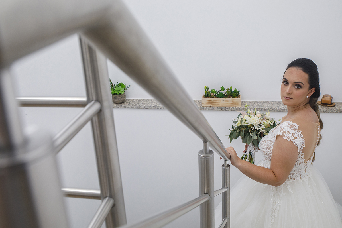 Através da perspetiva de uma lente encostada ao corrimão, vemos Marta lá em baixo, de perfil, com uma mão apoiada no ferro e a outra a segurar o bouquet enquanto olha para a câmara com um misto de timidez e doçura, envolta por luz e expectativa.