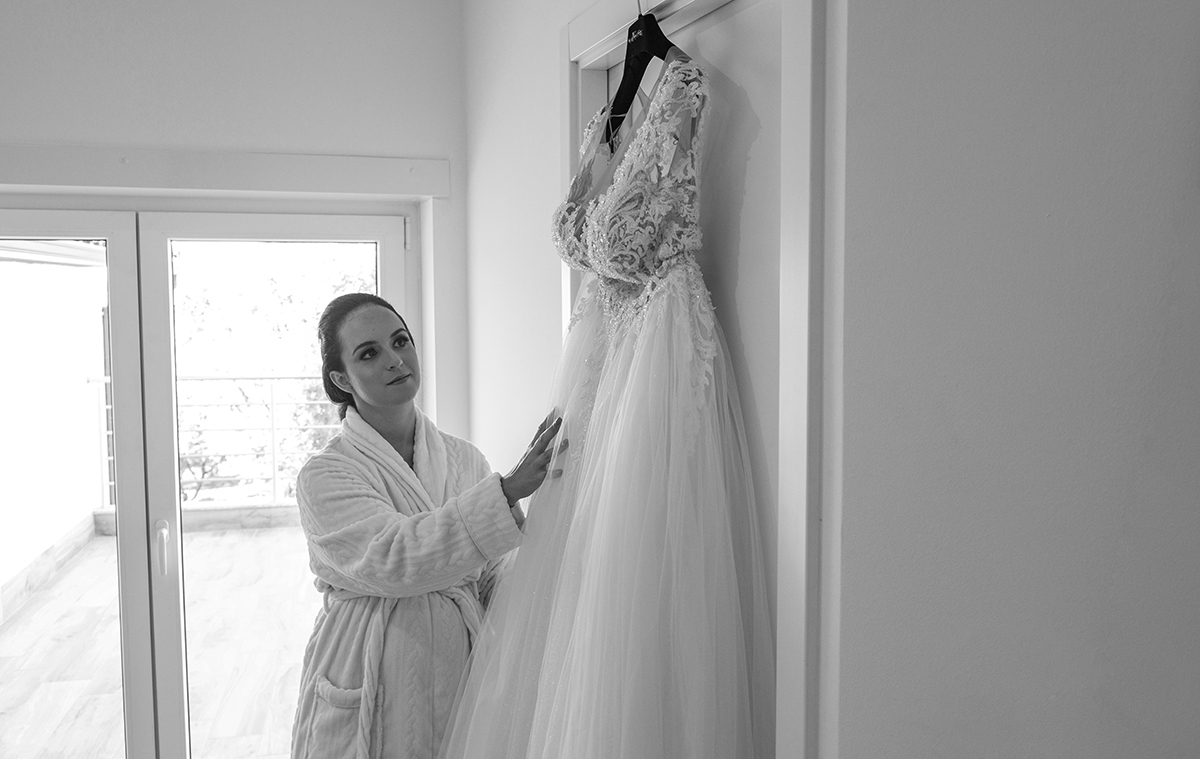 Em preto e branco, a noiva passa levemente a mão pelo vestido que repousa numa porta, num gesto de carinho e contemplação que revela o amor que sente pelo dia que está a nascer.